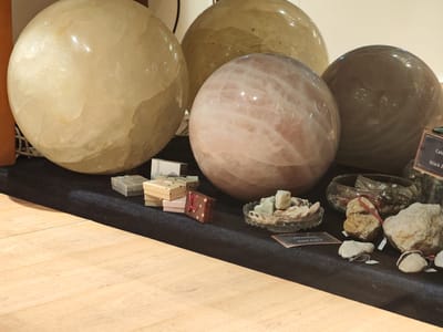 The image displays a collection of large, polished spheres and raw stones arranged on a black felt surface. In the foreground, a light-colored wooden surface is visible. The spheres vary in color and translucency, with one large, pale yellow sphere dominating the left side. To its right, a pinkish, marbled sphere is prominent, followed by a darker, grayish-brown sphere. Behind these, another large, pale yellow sphere is partially visible. Several small, gift-wrapped boxes, tied with ribbons, are stacked to the left of the pink sphere. To the right of the pink sphere, a small, glass bowl contains rough, light green stones. Further to the right, a larger glass bowl holds an assortment of colorful beads. In front of the glass bowls, several raw, irregularly shaped stones and pebbles are displayed, some with small white price tags attached. A dark, rectangular sign with text is partially visible in the lower right corner. The background is out of focus but appears to be a light-colored wall or display. This scene is likely from a shop or market stall, possibly selling geological specimens or decorative items. The mention of Schwangau, Germany suggests this is a retail environment located in or near that town.