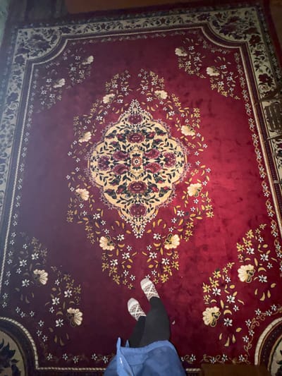 Indoors in Dehqonbozor, Uzbekistan, a large, rectangular deep red rug covers the floor, viewed from an overhead perspective. The rug features an ornate floral design with a prominent central medallion in cream and gold tones, detailed with red and dark red floral patterns. Scattered white, yellow, and beige floral sprays with green foliage are distributed across the red field. A multi-layered border in cream and darker tones frames the rug. In the lower-center of the frame, the lower legs and feet of an individual are visible. The individual wears black trousers or leggings, white socks with a light brown spotted pattern, and a portion of a blue garment is visible over their lap. The feet are positioned at an angle on the rug.