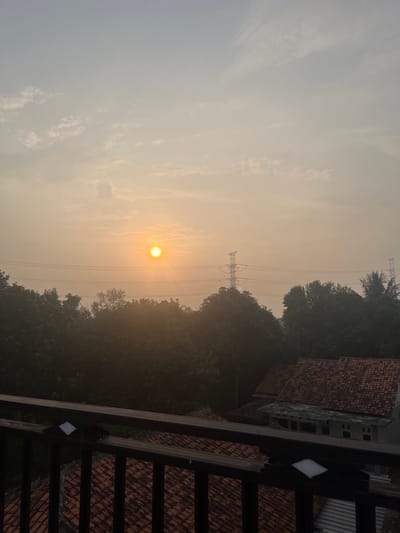 A hazy, muted orange sun is visible in the upper left quadrant of the image, partially obscured by thin, wispy clouds. The sun is casting a soft glow over the scene, which appears to be a residential area with a dense line of trees in the middle ground. Behind the trees, two tall electricity pylons with numerous power lines are visible against the sky. In the foreground, a dark railing with vertical supports runs horizontally across the lower portion of the frame, obscuring a view of several houses with terracotta tile roofs. The overall atmosphere is serene and hazy, possibly indicating early morning or late afternoon light with some atmospheric moisture. The location is identified as Pasir Gadung, Indonesia.