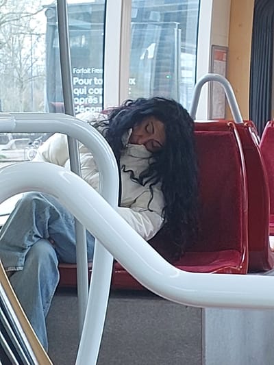 The scene depicts the interior of a public transport vehicle, consistent with a tram, located within the city of Illkirch-Graffenstaden, France. A lone female individual with long, dark, curly hair is shown in a state of sleep or unconsciousness, slumped forward on a red upholstered seat. She is wearing a light-colored, possibly white, puffer jacket and light blue jeans, with her head resting against her left arm, obscuring her face. White, curved metal handrails are positioned in the immediate foreground, partially obstructing the view of her lower body and the vehicle's floor. Through the large window on the left, a blurred daytime outdoor environment is visible, featuring deciduous trees with bare branches and a French advertisement sign partially legible as "Forfait Free." The interior is illuminated by natural light.