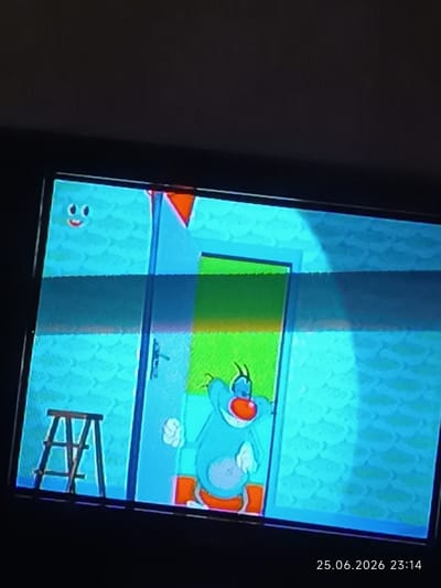 The image shows a television screen displaying a cartoon scene. The main subject is a blue cartoon character, likely Oggy from "Oggy and the Cockroaches," standing in a doorway. The character has large eyes, a red nose, and is wearing orange shoes. To the left of the character, there is a small wooden step stool. The background is a light blue, textured wall. There is a green rectangle visible within the doorway. The lighting suggests an indoor setting.

In the upper left corner of the screen, there is a smaller blue cartoon face with wide eyes and a smile. The screen itself shows some digital distortion, with horizontal lines of color banding across the image.

At the bottom right of the television screen, there is a timestamp: "25.06.2026 23:14". The location context provided is Bolhrad, Ukraine. The time and date stamp suggest this is a recording or a still image captured from a television broadcast. The overall mood of the cartoon appears lighthearted.