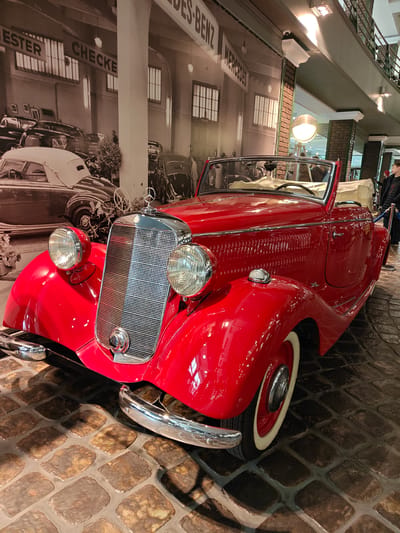 The image shows a red vintage Mercedes-Benz convertible displayed on a cobblestone floor. The car has a polished chrome grille and whitewall tires. Behind the car is a black and white mural depicting a Mercedes-Benz dealership or garage, with "ESTER CHECKER" and "MERCEDES-BENZ" visible on the building. A large industrial-style lamp is positioned near the car. A person is standing in the background, near a blue rope or barrier. The scene appears to be inside a museum or exhibition space. The location mentioned, Архангельское, Russia, might be where the car is displayed, although not directly visible in the image.
