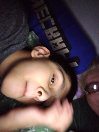 The image is a low-resolution, blurry selfie taken indoors. Two individuals are partially visible. The primary subject is a person lying down, facing the camera, with their head tilted. Their right ear and part of their face, including an eye and nose, are in focus. They are wearing a gray shirt. Behind them, a blue pillow with a "Minecraft" logo is visible. To the right of the primary subject, another person's face is visible, also at an angle, wearing glasses. The background appears to be a bed or similar soft surface. There is no indication of the city of Al Mas'oudi, United Arab Emirates, in the image.