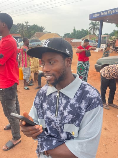 A medium-angle shot of a man in a black baseball cap and a unique button-down shirt holding a black smartphone. The man is smiling and appears to be looking at the phone. Several other individuals are visible in the background, some wearing brightly colored clothing and traditional patterns. The location seems to be a bustling area with vehicles and buildings in the distance. A sign that says "Adam-Tony Nigeria" is visible. The ground appears to be a dirt or clay surface.