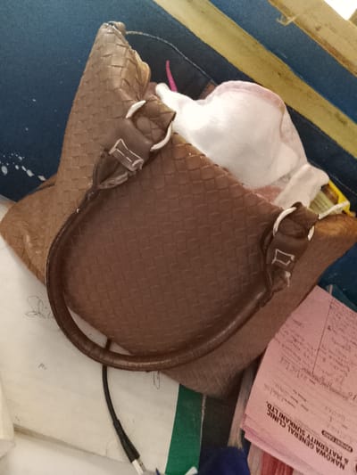 The image shows a brown woven handbag resting on top of other items, including a pink medical form. The handbag has a curved handle and appears to be made of leather. A white garment, possibly a bra or undergarment, is partially visible inside the handbag. A black cable, possibly a phone charger, is also lying near the handbag.

The pink form is from NAKOWA GENERAL CLINIC & MATERNITY SUNKANI LTD., located in Sunkani, Nigeria. The form contains handwritten text, likely medical information or patient details. There are also some smudges and marks on the paper, suggesting it has been handled.

The background consists of a striped surface, with alternating bands of blue and yellow. This could be part of a piece of furniture or a textured wall. The overall scene appears to be indoors, with a cluttered or unorganized arrangement of personal belongings. No people are visible in the image. The lighting suggests it is daytime, but the specific time of day or weather conditions cannot be determined. The emotional tone is neutral, focusing on the objects and their arrangement.