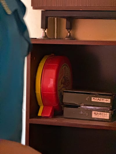 The image shows two shelves of a wooden cabinet. On the top shelf, there is a red and yellow circular object. On the bottom shelf, there are two green boxes with labels. The left box has a label that reads "ПЛАСТИК" (Plastic). The right box has a label that reads "ШАРЕЫ" (Balls) and "ПЛАСТИК" (Plastic). A person wearing a blue garment is partially visible on the left side of the image. The background is a wall. The scene appears to be indoors, possibly a storage area or a room in Naro-Fominsk, Russia, given the Cyrillic text on the labels.