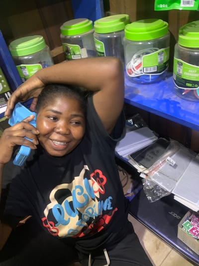 A woman smiles while holding a blue, boxy object to her ear, presumably mimicking a phone. She lies next to a blue shelf filled with plastic containers of phone chargers. An Acer laptop and other electronics are visible near her. She wears a black t-shirt with a large, colorful graphic design. The scene appears to be indoors, possibly in a shop or storage area.