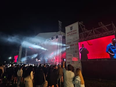 This image depicts an outdoor night concert or festival taking place in Elin Pelin, Bulgaria. The central focus is a brightly lit stage under a dark sky, featuring a truss structure and an overhead canopy. On stage, at least three performers are visible: two vocalists (one in a light shirt, one in a darker shirt) and a DJ or musician positioned behind equipment. Intense stage lights emit white and red beams through atmospheric haze, illuminating the performers and the immediate stage area.

To the right of the stage, a large, vertically oriented white banner prominently displays "ШОПСКИ ПРАЗНИК" (Shopska Praznik), indicating a local festival, along with numerous sponsor logos such as FLYNET, ECOPETROL, NOBEL, Semmelrock, AEC-X, BRAMAC, MEГАРИ, Poбoно, Fresh, Market, and Baceнa. Flanking the stage are two large LED video screens. The screen on the right predominantly displays a man in a blue shirt, likely one of the performers, against a bright red background. A similar screen to the left, partially visible, shows another performer, silhouetted against a red background.

In the foreground and midground, a substantial crowd of people, predominantly young adults, faces the stage. They are casually dressed. Several individuals in the crowd are holding up smartphones, suggesting they are recording or photographing the event. The ground appears to be an open grassy area. The overall scene is vibrant with strong artificial lighting from the stage and video screens, contrasting sharply with the dark night sky.