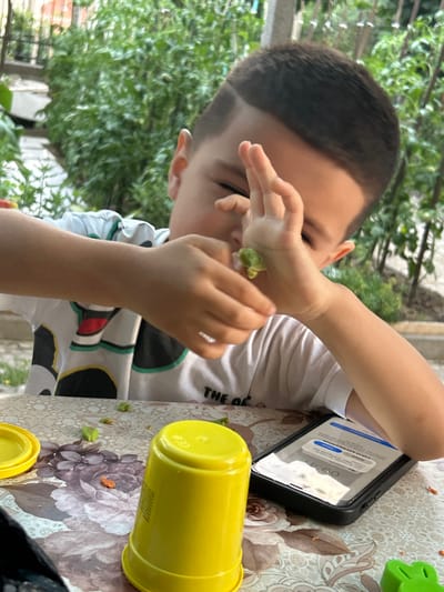 In Sveti Vlas, Bulgaria, a young boy is seen from the chest up, in the foreground, at a table outdoors. He is wearing a white t-shirt with a colorful pattern and is holding a small green object, possibly food, in his left hand, bringing it towards his face. A yellow cup is prominently placed on the table, to the left of the boy, and a smartphone is lying face up in front of him. The table surface has a floral pattern. In the background, lush green foliage suggests an outdoor setting, possibly a garden or yard, with some hints of urban structures behind the greenery. The lighting indicates it is daytime.