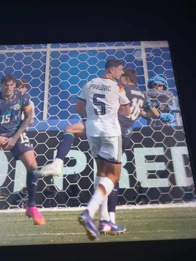 The image shows a soccer match with players near a net. One player in a white jersey with the name "PAVLOVIC" and the number "5" is visible. Another player in a dark blue jersey with the number "15" is also present. The background features a hexagonal net. The scene appears to be a stadium, potentially the Signal Iduna Park in Dortmund, Germany, given the context of a soccer match. The players are in action, with one appearing to kick a ball or be in the process of a play. Other individuals, possibly spectators or team personnel, are visible behind the net.