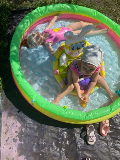 Two young girls are enjoying a sunny day in an inflatable rainbow-colored kiddie pool. The pool is filled with clear water and has a rabbit-shaped inflatable toy in it. One girl, wearing a pink swimsuit, is lying on her back in the water, her eyes closed, appearing relaxed. The other girl, wearing a leopard-print swimsuit and a light gray cap, is sitting upright on the inflatable toy, holding a yellow rubber duck. The scene is set outdoors on a grassy area next to a black tarp. Several pairs of children's shoes are placed on the tarp near the pool. The image conveys a sense of fun and summer enjoyment. The time appears to be daytime, with bright sunlight casting shadows. The location is specified as Marinopil, Ukraine. No visible text is present in the image.