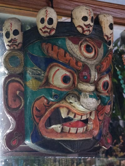 This media file features a close-up, frontal view of a colorful, carved wooden mask. The mask is designed to represent a fierce, mythical creature, possibly a deity or demon from Tibetan or Himachali folklore. It is adorned with multiple smaller, skull-like representations at the top, each with simple black eye sockets and red markings around the mouth.
The main mask has an elaborately carved face with wide, bulging eyes, some with red outlines and black pupils. The mouth is open in a terrifying grimace, revealing sharp white teeth and a red interior. The mask is painted in a vibrant palette of colors, including teal, orange, red, blue, yellow, and white, with intricate patterns and textures. There are blue gemstone-like accents near the eyes and on decorative elements. Flanking the main face are stylized ear-like structures, also carved and painted. The overall impression is one of power and intensity, common in masks used in traditional religious dances or performances in regions like Manali, India.
The setting appears to be indoors, with a reflective surface in the foreground, possibly glass or polished wood, suggesting the mask might be displayed on a shelf or as part of a collection. In the background, blurred greenery and a hint of beaded decoration can be seen, adding to the exotic and cultural context. The lighting is soft and diffused, highlighting the details of the mask. No visible text is present in the image. The mask itself evokes a sense of awe and perhaps a touch of fear, reflecting its cultural significance in representing powerful spiritual or mythical entities.