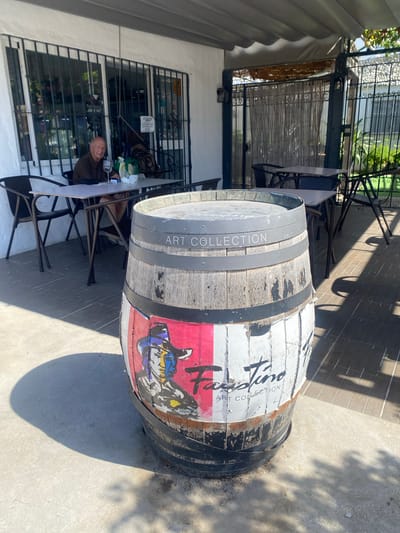 A barrel with a colorful artistic design, possibly advertising "Hauterive Art Collection," is placed on a patio. The barrel features a prominent logo or illustration on its side, with weathered wooden staves and dark metal bands. The scene appears to be outdoors, with a tiled patio floor casting shadows from unseen trees or structures, suggesting a sunny day.
To the left of the barrel, a white table with chairs is set up. A man is seated at the table, wearing a brown shirt and looking towards the viewer. He appears relaxed, with a glass on the table in front of him. Behind the table, a building with large windows is visible. These windows are covered by decorative metal grates, and through them, interior displays of what appears to be a shop or gallery can be seen. The building has a white facade.
The overall setting suggests an outdoor seating area of a business, possibly a cafe, restaurant, or gallery, located in Estepona, Spain, given the provided location context. The presence of the barrel and the artwork implies a creative or artisanal establishment. The lighting indicates it is daytime, likely with clear, sunny weather, as evidenced by the strong shadows.