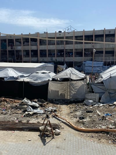 An outdoor scene in Jabalia, Palestinian Territory, captured during daylight under a clear blue sky with scattered white clouds. The foreground features an unpaved, rubble-strewn dirt ground that transitions to a paved surface at the bottom. A pile of rusty rebar lies on the left foreground. A brown tripod stands centrally, stabilized by rocks at its base, casting a shadow. An orange-brown, flattened hose or pipe extends across the dirt from left to right. The midground contains numerous makeshift shelters and tents constructed from gray, white, and tan tarpaulins and fabric sheets, arranged amidst additional debris. Several small figures, possibly children or adults, are visible near the right side of these shelters. The background is dominated by a multi-story concrete building, likely a damaged residential structure, with numerous open window or balcony frames. Laundry hangs from several balconies. A black water tank is visible on the roof, and exposed structural elements or damaged scaffolding are present on the rooftop. A faint emblem or text is discernable on the building's lower-left section.
