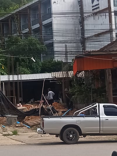 The image shows a construction site in Ban Ao Nang, Thailand. A silver Nissan pickup truck is parked on the street in front of a partially constructed building with exposed rebar. Two men are visible within the construction zone; one is wearing a light-colored shirt and is standing, while the other is wearing a hat. A black tarp is partially covering the construction area. In the background, a multi-story building is visible, with electrical wires running across the frame. The environment appears urban, with signs of ongoing development or renovation.