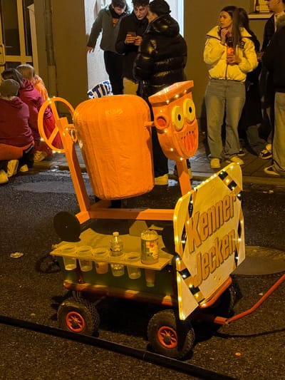 A nighttime scene depicts a custom-built, wheeled contraption on a dark asphalt surface, with a group of approximately ten individuals standing in the background against a light-colored building wall in Wasserbillig, Luxembourg.

The primary subject is a mobile, orange-colored structure resembling a stylized concrete mixer with an attached sign. It features a large, horizontally oriented cylindrical drum, and a separate, smaller component to its right that depicts a cartoonish face with prominent eyes and a large, jagged mouth. Both elements are adorned with small, warm-white LED lights. This structure is mounted on a chassis with four black wheels featuring orange-red hubs. A wooden shelf on the contraption's lower section holds six clear plastic cups, a clear plastic bottle of water, and a plastic bottle containing orange-colored liquid. A red hose extends from the contraption's lower right side onto the ground. Attached to the front-right of the contraption is a yellow and black striped sign, also illuminated with small white LED lights, bearing the text "Kenner Jecken". Debris, possibly a flattened silver can, is visible on the asphalt to the left of the contraption. A circular manhole cover is partially visible to the right.

In the background, positioned in an apparent building entrance or recessed area, multiple individuals are present. To the far left, two partially visible individuals are crouched or seated, one in maroon, another in orange. Three individuals stand centrally: one male in a dark greenish-grey hooded jacket looking down at a smartphone, a male with dark hair and glasses also viewing a smartphone, and a female in a shiny black puffer jacket standing close by. To their right, a female with long dark hair, wearing a bright yellow puffer jacket and light blue jeans, holds a clear drinking container to her mouth. Other individuals are partially visible further to the right. The scene is illuminated by ambient artificial light sources, casting shadows.