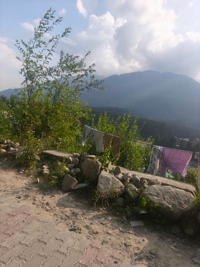 The image depicts a serene landscape scene in Manali, India, likely in the late afternoon given the soft, angled sunlight. In the foreground, a dirt path paved with interlocking stones leads towards the right. Large, weathered rocks and lush green vegetation line the path. A clothesline strung with several items of laundry, including a light blue garment, a brown cloth, and a purple sheet, is visible stretched across the midground. Behind the clothesline, dense green foliage creates a natural backdrop, with a distant, mist-shrouded mountain range dominating the horizon. The sky is a mix of bright blue and scattered, fluffy white clouds, indicating fair weather. The overall impression is one of peaceful rural life, with everyday activities like laundry drying set against a majestic natural backdrop. There is no visible text in the image.