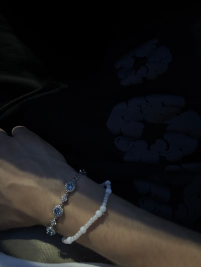The image shows a close-up of a person's left arm and hand, adorned with two bracelets. The bracelet closest to the hand is made of small, white beads, while the bracelet further up the arm is a silver chain with three light blue gemstones and small diamond-like accents. The background is dark, with what appears to be a black garment with a faint, light-colored floral or cloud-like pattern. The lighting suggests an indoor setting, possibly at night. There is no visible indication of the city of Vaughan, Canada in the image.