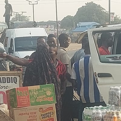 This image depicts a bustling outdoor scene in Gudi, Nigeria, characterized by a hazy atmosphere under a pale sky. In the foreground, a group of individuals stands around roadside stalls and vehicles. A woman wearing a dark, patterned head covering and garment is prominently positioned in the lower left, with a child in a red and white striped shirt standing adjacent to her. Behind them, a man extends his left arm, pointing or gesturing toward the left side of the frame.
To the left, a white passenger van has a man standing on its roof, adjacent to strapped cargo bundles. In the middle ground, several men are standing, observing various directions. To the right, a smaller white vehicle has its front passenger door open, revealing a man in a red shirt in the driver's seat. Another man, wearing a blue and white striped shirt, stands with his back to the viewer near this vehicle.
Merchandise is visible in the lower portion of the frame: a wooden stall displays the partial word "ADDIQ" and is stacked with cardboard boxes of "Viva Plus" and "Three Crowns Instant Filled Milk Powder." To the right, another display features numerous plastic bottles of water and other beverages. The background shows distant buildings, green trees, and utility poles with overhead lines.