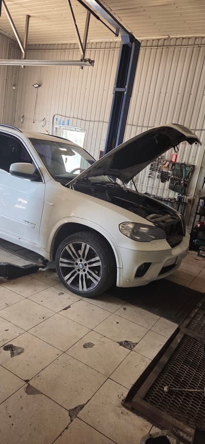 An interior shot captures a white BMW SUV positioned on a car lift in a garage. The vehicle's hood is open, revealing the engine compartment. The garage has corrugated metal walls and a tiled floor. Tools are visible on a rack in the background.