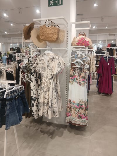The image shows a retail store interior, likely a clothing boutique, with racks of apparel and accessories. In the foreground, a white rack displays denim skirts and some patterned shirts. To the right, a dress with a floral print hangs on a hanger. Further back, more clothing racks are visible, featuring various garments like dresses, tops, and pants, along with straw bags and hats displayed on shelves. The store has a modern, clean aesthetic with white shelving and light-colored flooring. The lighting appears to be artificial, coming from ceiling fixtures. An illuminated green exit sign with an arrow is visible on the upper left portion of a white pillar. The general atmosphere suggests a bright and organized shopping environment. This scene could be located in Ávila, Spain, a city known for its well-preserved medieval walls and historic architecture, which might contrast with the modern retail space depicted.