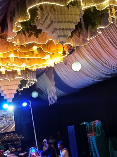 The image depicts a lively indoor or covered outdoor event taking place at night, likely in Pangalengan, Indonesia. The scene is dominated by elaborate ceiling decorations and lighting. Multiple tiers of sheer white and gold fabric, interwoven with artificial greenery, are illuminated by warm, glowing lights, creating a decorative canopy overhead. Additional white and blue fabric drapes are suspended across the ceiling, from which hang white spherical lights.
In the lower portion of the image, a stage or performance area is visible, brightly lit by blue spotlights. A group of people, primarily young men, are gathered on the stage. One person in the foreground is actively playing a red and white kendang, a traditional Indonesian drum, indicating a musical performance is underway. Other individuals are standing or sitting behind him. To the far right, a stack of colorful plastic chairs (green, red, and blue) suggests a seating area for attendees.
A white banner or sign is partially visible on the left side of the stage. Readable fragments of text on the banner include "JANG", "Bandung", "SINGGAH DI", "KAYANG DI", and "GEDUNG". These likely pertain to the event's name, location, or organizers, with "Bandung" providing a regional context to the location of Pangalengan. The overall ambiance is festive and cultural, characteristic of an evening gathering or celebration.