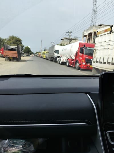 The image captures a traffic jam in Tingkeummanyang, Indonesia, viewed from the interior of a car. A line of trucks stretches down a paved road, suggesting a disruption in traffic flow. The foreground shows the dashboard of the car, with a digital display on the right. In the middle ground, a variety of trucks are visible, including a red tanker truck with "PT GUNUNG" written on it, and several other trucks of various colors and sizes, some covered with tarps. The background reveals buildings, utility poles, and trees lining the roadside. The sky is overcast.