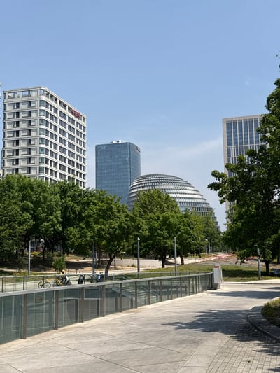 The image captures an outdoor scene in Chaoyang District, China, under a clear, blue sky. Prominent modern architectural structures, including several high-rise buildings with distinctive designs, dominate the background. The buildings feature a mix of straight lines and curvilinear forms, with glass and steel facades. Lush green trees partially obscure some of the buildings.
A pathway stretches along the right side of the frame, featuring a gray concrete surface and a glass barrier. Bicycles are parked near the glass barrier. The pathway transitions into a walkway made of brick.