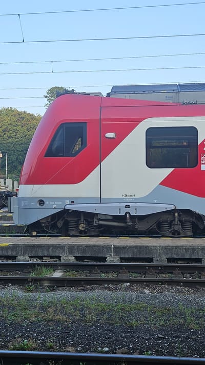 The image captures a side view of a red and white passenger train, identified by the "ÖBB KOSICE" marking on its side, at the Kysak, Slovakia train station. The train's locomotive, with its characteristic red front and white stripe, is prominently featured. The exterior of the train shows two windows, one on the front cab and another on the passenger section. Beneath the train, its wheels and suspension system are visible, resting on railway tracks. The station platform is a concrete structure, with gravel and vegetation around the tracks. In the background, trees and overhead power lines indicate an outdoor setting. The sky is clear and blue. There are no people visible in the image. The scene appears to be a static view of the train at the station, suggesting a pause in its journey.