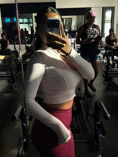 A woman takes a mirror selfie in what appears to be a gym or fitness studio in Long Beach, United States of America. She is the central subject, holding a smartphone with a reflective case to capture her image. Her face is mostly obscured by the phone. She has dark hair, styled sleekly, and wears a white long-sleeved crop top with a ribbed texture and burgundy or maroon high-waisted leggings. Her left hand, holding the phone, displays nails with what looks like a white French tip design and a ring on her ring finger.
The setting is an indoor fitness area, characterized by large mirrors and gym equipment. Reflected in the mirror behind the main subject are other individuals and exercise machines. One prominent reflection shows a person wearing a dark tank top with white text that reads "FUCK LOVE" and a pink baseball cap. Another reflection shows someone sitting on a piece of gym equipment. The equipment visible includes stationary bikes and resistance training apparatus with handles and bands. The lighting suggests daytime, with some natural light entering the space, creating reflections and highlights on surfaces. The overall atmosphere is that of an active workout environment.