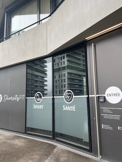 The image, rotated 90 degrees clockwise, captures the modern entrance and signage of a fitness center named "Diversity FIT" in Meyrin, Switzerland. The upper section of the entrance features a dark grey panel with white text and graphics. A prominent white circle with the word "ENTRÉE" (Entrance) is visible on the left. To its right, detailed information is listed, including "ENTRAINEMENTS FONCTIONNELS ET PRÉPARATION" with "Cours privés", "Cours semi-privés", and "Cours en petit groupe". Below this, "ENTRAINEMENTS MÉTHODE NOVAMUM" is mentioned, tailored for "femmes, pendant la grossesse et la maternité". Contact details include "@diversity.fit", "www.diversityfit.ch", and the phone number "+41 79 723 56 92".

Below this panel, two large glass window panes display further signage. The upper window features "SANTÉ" (Health) next to an icon of a heart with an ECG line, while the lower window shows "SPORT" alongside an icon depicting a person lifting a dumbbell. The "Diversity FIT" logo is also present in the lower left corner of these windows. The glass reflects a tall, multi-story building with numerous windows, indicating an urban setting and likely daytime conditions. An overhead canopy, with a textured, light-colored underside and integrated strip lighting, extends over the entrance. No individuals or active events are depicted; the scene is a static architectural shot of the fitness center's facade.