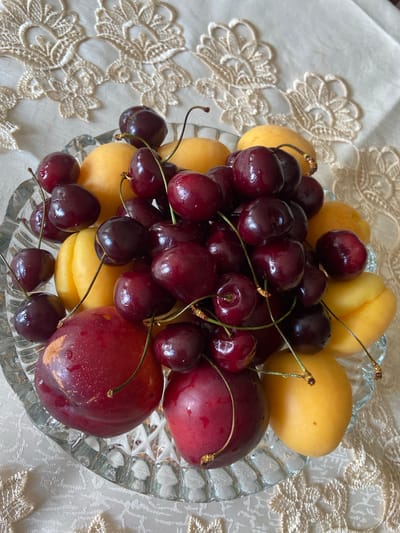 A clear glass bowl filled with ripe cherries and apricots is placed on a white tablecloth with intricate lace trim. Two red plums are also visible in the bowl. The fruits are wet, suggesting they have been recently washed. The lighting indicates it is daytime, with a soft light source coming from above. The image was taken in Askarovo, Russia.