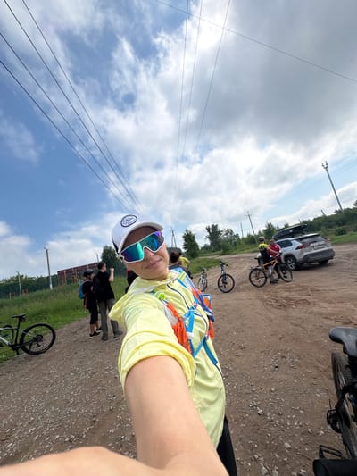 In Kemul, Russia, on a gravel road under a cloudy sky, a group of people with bicycles are gathered. A woman in the foreground, wearing a yellow jacket and reflective sunglasses, takes a selfie. Behind her, several individuals are standing near bicycles. A silver SUV with a roof rack is parked nearby, and trees and a fence are visible in the background. Overhead power lines stretch across the sky. The scene suggests a cycling outing or event in a rural or semi-rural area.