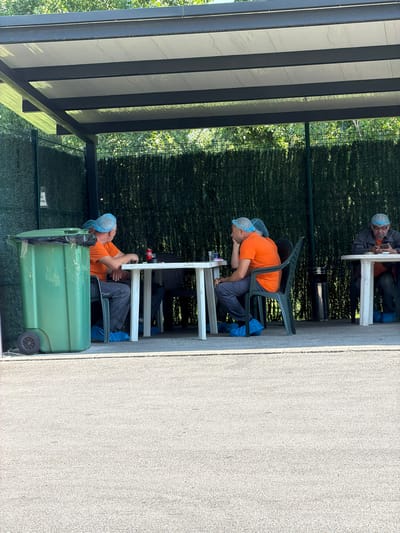 The image depicts an outdoor seating area under a covered structure. Three individuals are seated at tables, all wearing blue hairnets and blue shoe covers. Two individuals in orange t-shirts are seated at a white table in the foreground, facing away from the camera. One of them is looking at his phone, while the other appears to be looking forward. A third individual, wearing a dark shirt, is seated at a table further back on the right, also looking at his phone. A green trash bin is visible to the left of the image, next to the covered area. The background consists of a dense green hedge and trees. The ground is paved with a light-colored, textured material. The setting appears to be Lozen, Bulgaria, possibly a break area at a facility.