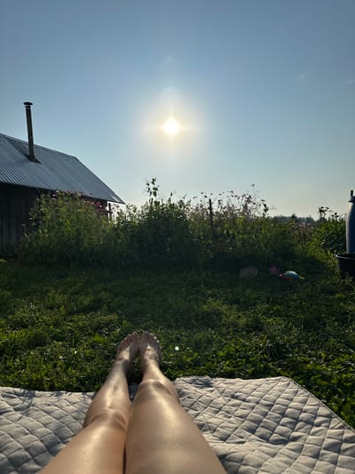 The image depicts a person's legs and feet resting on a quilted blanket laid out on a grassy area. The perspective is from the person's point of view, looking forward towards a clear blue sky with a bright sun. In the background, there is a rustic wooden building with a metal roof and a chimney, situated behind lush green vegetation and some pink flowers. Further in the distance, more greenery and trees are visible under the sunny sky. The overall scene suggests an outdoor, rural setting, possibly a backyard or a field, in the village of Sredneye Kechevo, Russia. There are no other people visible in the frame.