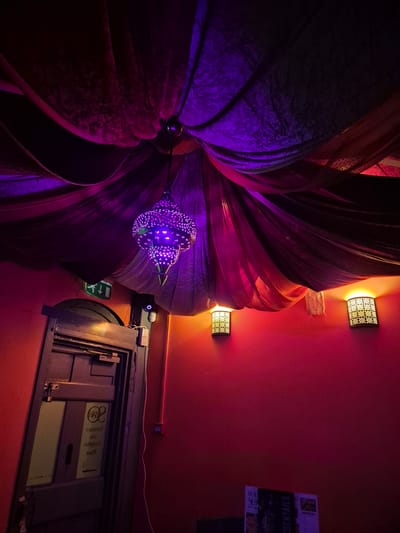 The image, taken from a low angle in what appears to be an interior space in West Suffolk, United Kingdom, shows a ceiling adorned with decorative elements. A large, flower-like fabric arrangement is the dominant feature, illuminated with purple and red lighting. Hanging beneath it is a smaller, decorative lamp with a patterned design, also lit in purple.

The ceiling is painted a dark red. Two decorative wall sconces with floral-patterned cutouts provide ambient lighting. To the right, there's an emergency exit sign with a green arrow pointing to the left. Below it is a framed display with text and images that appear to be promotional material, partially obscured by the darkness. There is also a door in the lower right corner.
