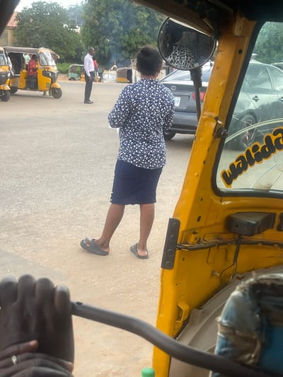 A medium shot shows a woman in a navy blue and white floral print top and a dark blue skirt, standing with her back to the camera in a dusty outdoor area. She is wearing grey slip-on sandals. To her left, a man in a white shirt and black pants stands facing away from the camera, and a yellow auto-rickshaw is parked. In the background, other auto-rickshaws and a silver sedan are visible. The foreground shows the interior of a yellow auto-rickshaw, with a dark handle bar and the hand of the person operating it visible. The scene appears to be a street or transport hub in Galadimawa Village, Nigeria, characterized by unpaved ground and several vehicles.