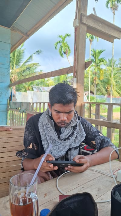 A young man with dark hair and a mustache, wearing a black shirt with a patterned scarf around his neck, is seated at a wooden table. He is looking down and holding a smartphone in his hands, with a white charging cable connected to it. To his left, a glass mug filled with a dark beverage and a straw sits on the table. In the background, palm trees and a cloudy sky are visible, suggesting an outdoor setting, likely in Grong-grong, Indonesia, characterized by a rustic, open-air structure with wooden beams and light blue siding. The man's posture and focus on the phone indicate he is engaged in using the device.