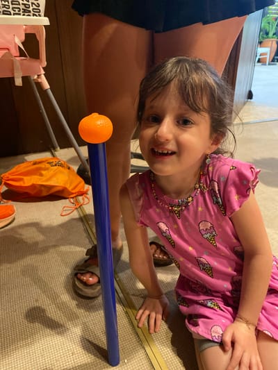 A young girl wearing a pink shirt with ice cream cones and stars is on a beige carpet with a blue tube with an orange ball at the end. There is a pink toy and an orange bag with drawstrings in the image. A woman in a black skirt and brown sandals is partially visible on the left side of the frame. The location is Marina Maroma, Mexico.