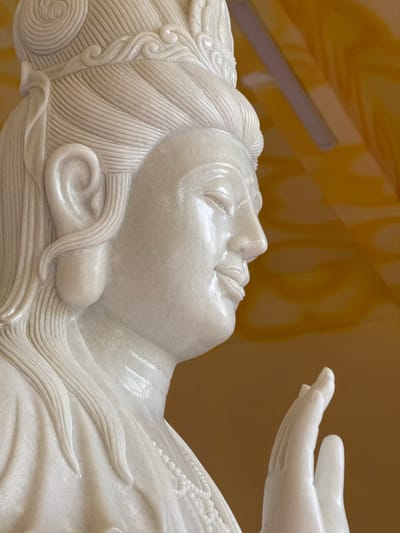 A highly polished white marble statue, depicting a deity or figure, is shown from the upper torso upwards, positioned within an interior setting in the city of Đăk Mar, Vietnam. The statue faces right in profile, featuring meticulously carved hair pulled back and adorned with an ornate crown. Its eyes are depicted as downcast, and a subtle smile is present on its lips. A necklace of sculpted beads rests on the figure's chest. The right hand is raised, palm facing forward with fingers extended and slightly curved. The immediate background consists of a uniformly golden-orange surface, against which a linear fluorescent light fixture is visible in the upper portion of the frame. The statue exhibits a smooth, reflective surface with fine granular textures.
