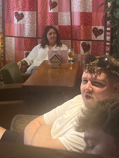 Two people sit at a wooden table. One person, closer to the camera, wears sunglasses atop their head and a white t-shirt. The other person, sitting across the table, also wears a white shirt. A beer glass, menu, and drink coasters are on the table. Behind them is a wall hanging with a red and white patchwork design featuring heart appliques. The image is rotated slightly, with the individuals' faces in the upper portion.