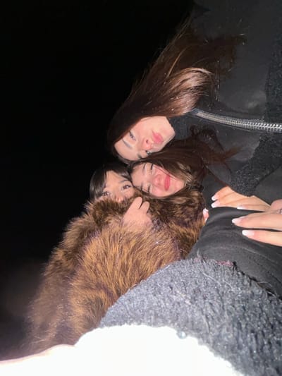 Three young women huddle closely together, faces illuminated against a dark backdrop. The woman on the left wears a black jacket and has long brown hair. The woman in the center also has brown hair and a slight smile. The woman on the right wears a brown fur jacket. Their hands are visible, indicating they are hugging. The background fades into darkness on the left and lightens slightly towards the right.