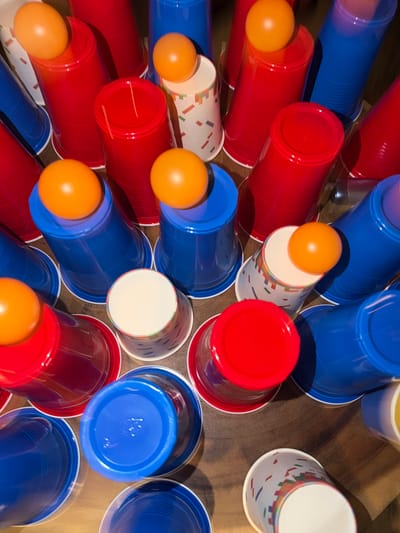 This is a top-down, close-up shot of a collection of red, blue, and white cups, many of which have orange ping pong balls resting on top. The cups are arranged in a scattered, somewhat haphazard manner on a wooden surface. Some cups are filled, others are empty, and some are patterned with confetti-like shapes. The lighting is somewhat dim, casting shadows that emphasize the texture of the cups and the wooden surface. There are no people visible in the image, and no specific geographical features to indicate a location in Heijen, Netherlands. The scene appears to be set up for a game of beer pong, a popular drinking game.
