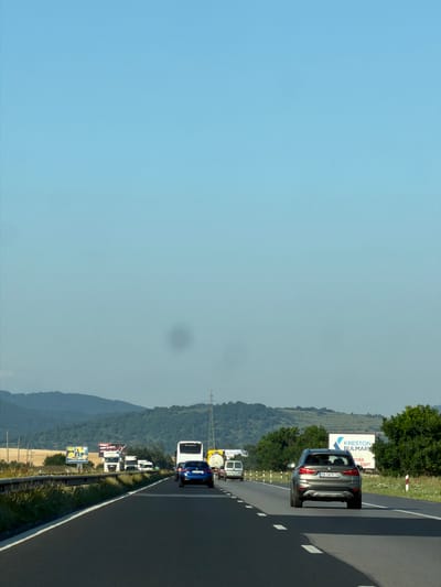 This media file shows a view from a vehicle traveling on a highway in Yana, Bulgaria. The scene is characterized by a clear, light blue sky and a lush green landscape on the left side of the road, suggesting a warm, possibly summer day.

The foreground is dominated by the asphalt of the highway, with white lane markings visible. A silver car is the most prominent object, positioned in the right lane. Behind it, a line of trucks and vans are seen traveling in the same direction. The vehicles are varied, including what appear to be commercial trucks and passenger vans.

In the background, rolling hills covered in dense green trees form a prominent feature. Several billboards are visible along the side of the highway. One billboard displays text that includes "KRESTON BULMARTA". Another billboard below it shows a picture of a person. The presence of a highway and billboards indicates a developed area, likely a common travel route.

The overall impression is one of a journey on a bright day, with the natural beauty of the Bulgarian landscape complementing the man-made infrastructure of the highway. There are no people prominently visible, and the focus is on the vehicles and the environment. The time of day appears to be daytime, given the bright sky.