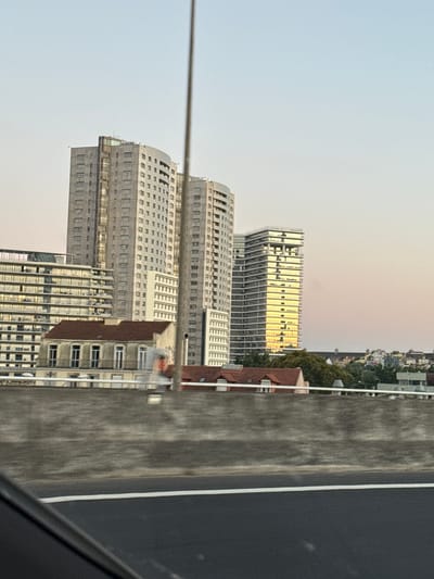 The image shows a view of modern buildings and a lower-rise building in the foreground, likely in an urban environment. In the background, a cityscape with trees and various architectural styles is visible under a clear sky with subtle hints of sunset colors. The overall scene is one of urban development with a mix of old and new structures. The image also shows a portion of a road and a concrete barrier, suggesting the vantage point is from a moving vehicle on a road or bridge. The image is a photograph of the city of Loures, Portugal, captured from an elevated perspective.