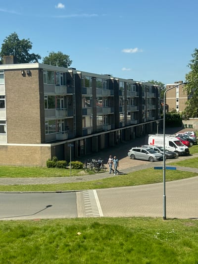 An exterior daytime shot shows a residential area in Breda, Netherlands. A long, multi-story, brick-faced building with repeating window patterns runs horizontally across the frame, receding into the distance. Multiple cars, primarily white, are parked adjacent to the building. A paved walkway and a narrow road run parallel to the building. Two individuals stand beside a group of parked bicycles. A lamppost is situated between the road and walkway. Green grass covers the ground on both sides of the walkway and road. The sky is blue with few clouds.