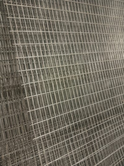 The image displays a tightly-cropped, close-up view of multiple sheets of metallic wire mesh, observed within the city of Kofar Ruwa, Nigeria, though no specific urban environment or geographical markers are visible. The primary subjects are numerous, uniformly constructed wire mesh panels, stacked horizontally, filling the entire frame. Each panel consists of thin, silver-grey metallic wires welded perpendicularly to form a consistent grid of square openings. The stacked arrangement results in a visual density on the left side of the frame where multiple layers overlap, making the underlying grids less distinct. Towards the right, the patterns of individual underlying mesh sheets are faintly discernible through the top layer, confirming the stacked nature of the material. No individuals, active subjects, or distinct background objects are present. The metallic surfaces reflect ambient light evenly, without casting discernible shadows.