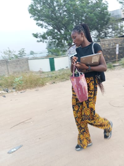 A woman walks along a dirt road holding a laptop and a pink shopping bag. She wears a black t-shirt, patterned yellow and black pants, and black shoes with buckles. Her hair is braided and pulled back. A piece of paper is in her mouth. In the background, there is a tree, a white building, and a stone wall. Debris lays on the ground near the woman.