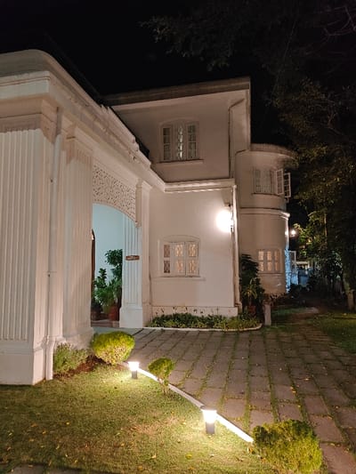 This image captures the illuminated exterior of a white, two-story colonial-style building at night, located in Kasa Kadavu, Mattancherry, Fort Kochi, India. The structure features a large entrance archway on the left with fluted white columns and intricate decorative carving above the arch. Inside the archway, potted plants are visible, along with a wooden sign reading "KASA KADAVU" and "STORAGE AREA". The building's main facade on the right side displays multiple multi-paned white windows; three are on the ground floor, and two on the upper floor, one of which is part of a rounded bay extension and is slightly ajar. The building is lit by several wall-mounted exterior light fixtures. In the foreground, a stone paver pathway runs alongside a manicured grass lawn, dotted with small, low-lying shrubs. Both the lawn and pathway are illuminated by multiple cylindrical ground-level lights. Dense dark foliage outlines the building against a dark night sky in the background. No individuals are present in the scene.