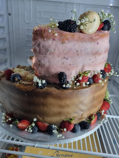 The image depicts a two-flavored cake, decorated with fresh berries and small white flowers, inside a refrigerator. The cake appears to be cut or partially eaten. One half is coated with pink frosting, while the other has a brown coating. Berries like strawberries, blueberries, and blackberries, along with the white flowers, garnish the top edges of the cake. A single macaron is placed on the pink side. The cake rests on a silver cake stand within the refrigerator, which features white wire shelves. No specific information about the location in Saarbrücken, Germany is discernible from the image.